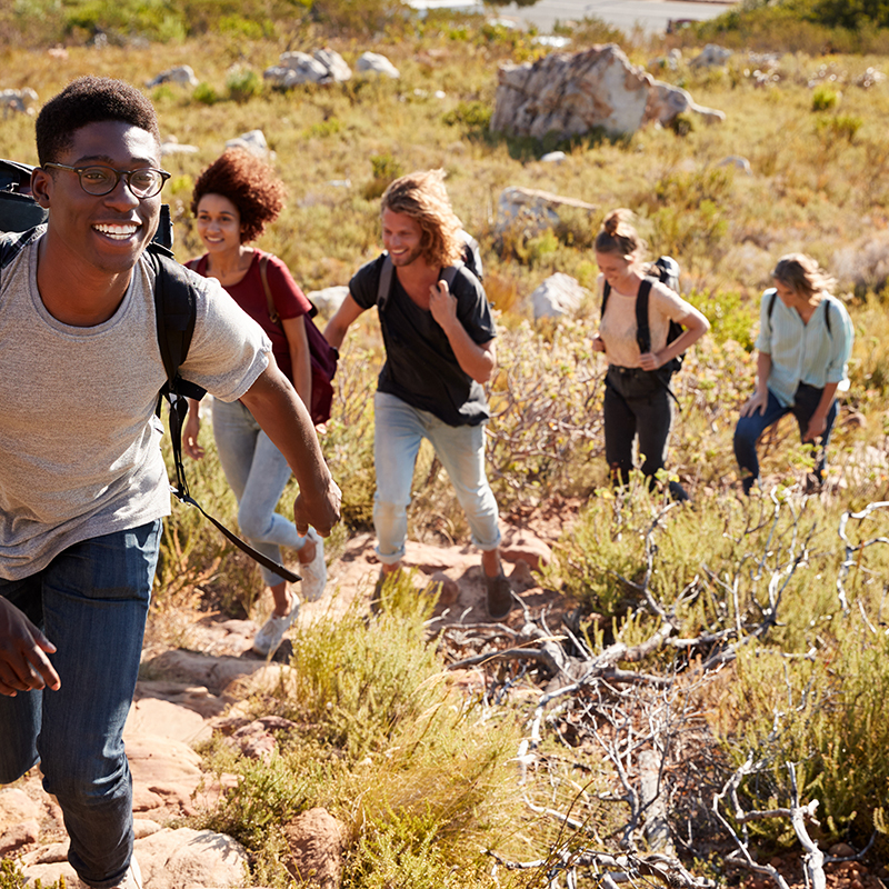 Millennial African American man leading friends hiking single file uphill on a path in countryside