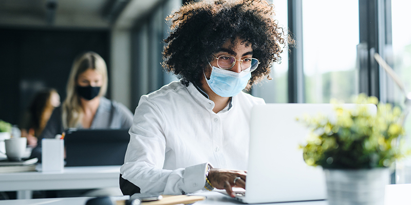 Portrait of young man with face mask back at work in office after lockdown, working.