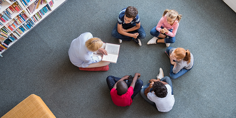 Teacher reading fairy tales to children sitting in a circle at library.