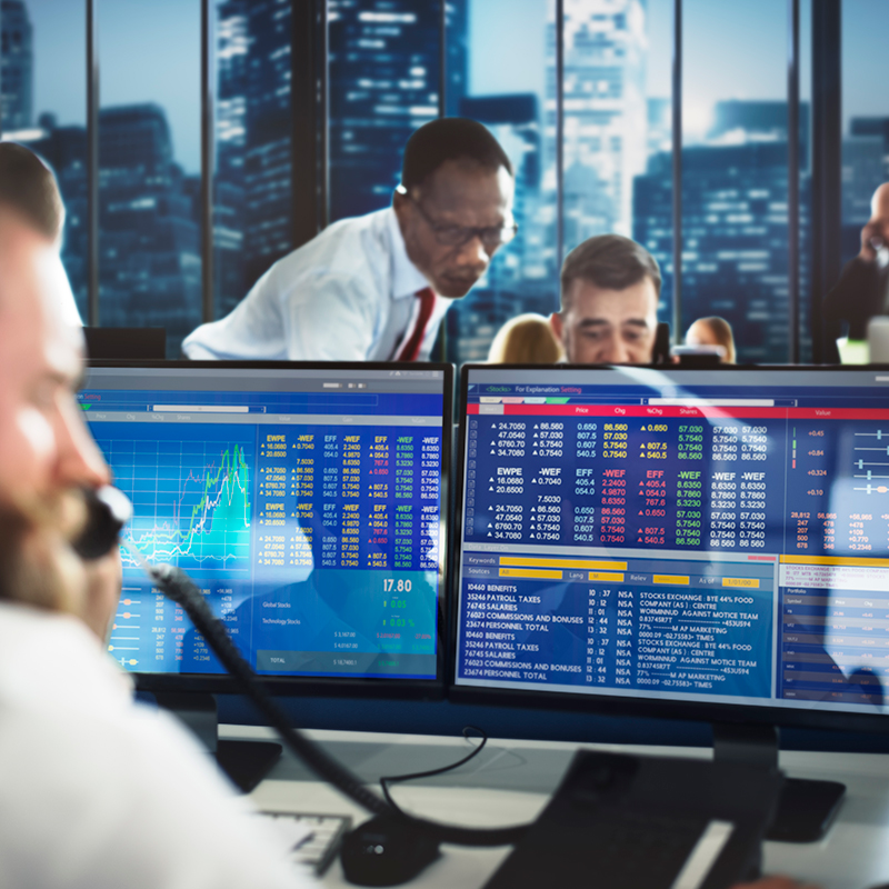 Two monitors showing financial info with two people in the background