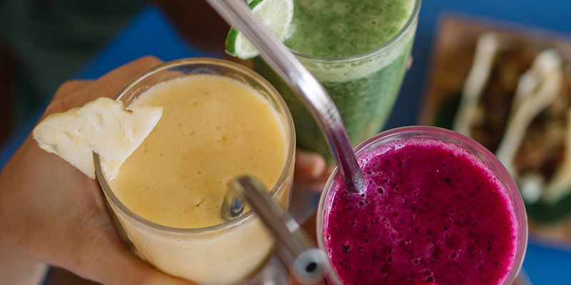 Cropped image of cafeteria customers holding smoothie cocktails and gesturing cheers during friendly meeting in bistro, selective focus on colorful milkshakes with mixed ripe fruits and fresh veggies