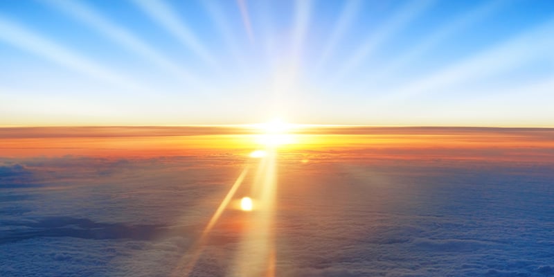 View on orange sunset, sun, blue sky, and ocean of clouds from airplane window.
