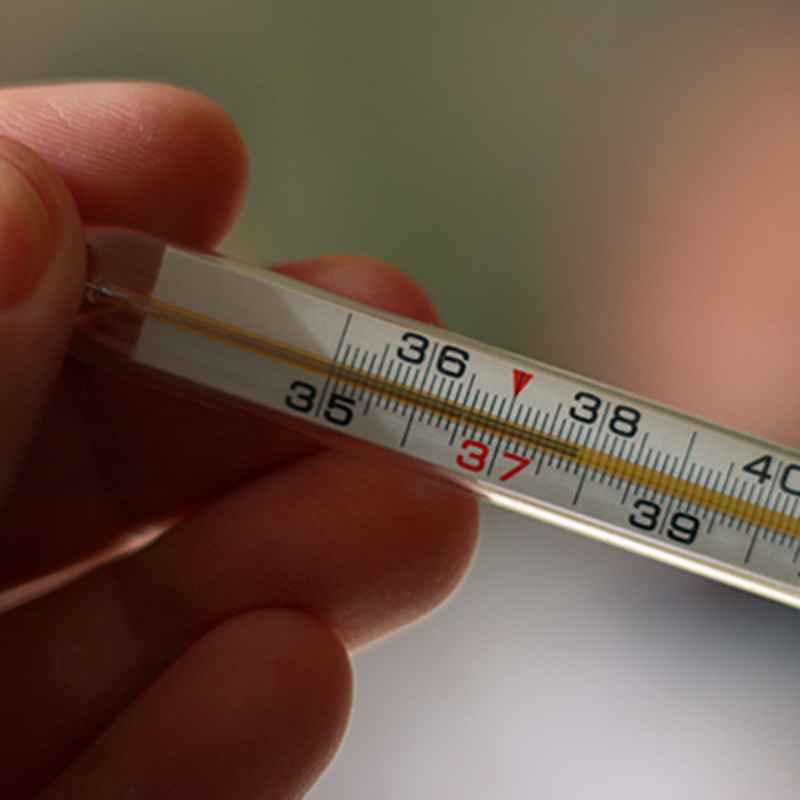 An alcohol thermometer shows a temperature of 37.9 on a blurred background of a sick girl lying on a bed